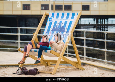 Aberystwyth Wales UK, mercoledì 26 settembre 2018 UK Meteo: due giovani donne seduto in una sedia sdraio gigante su Aberystwyth prmenade godendo di un pomeriggio di sole caldo sulla West Wales coast © Keith Morris / Alamy Live News Foto Stock