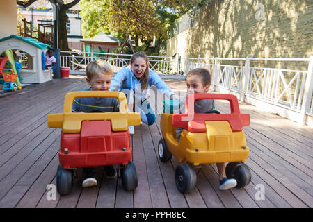 Due ragazzi di corsa in auto del pedale sulla terrazza in asilo nido curato da un insegnante Foto Stock