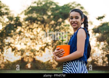 Basso angolo di visione di un sorridente ragazza tenendo un vaso di fiori in mano in piedi nel parco. Foto Stock