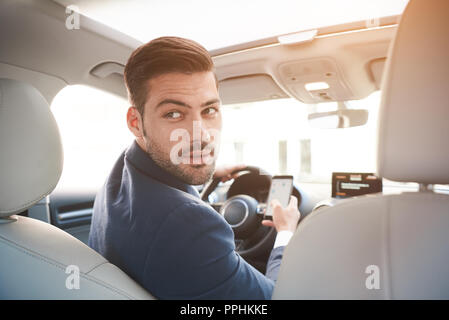 Ritratto di elegante imprenditore fiducioso in auto indossare tuta nera, girando per sedili posteriori con espressione seria. Foto Stock