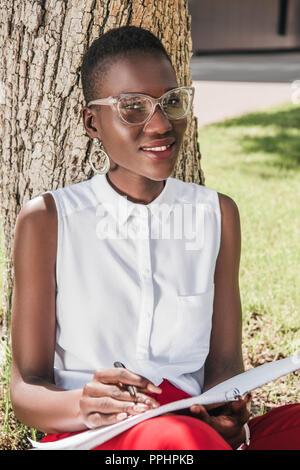 Elegante sorridente african american imprenditrice poggiando su albero nel parco e notebook di contenimento Foto Stock