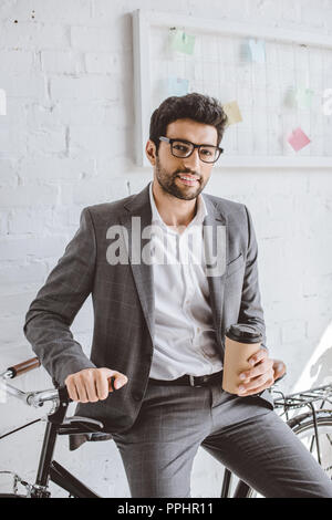 Sorridente imprenditore bello holding tazza di caffè e appoggiate su bici in ufficio Foto Stock