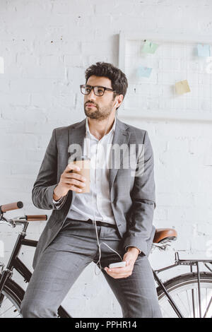 Imprenditore ascoltando musica con lo smartphone, tenendo tazza di caffè e appoggiate su bici in ufficio Foto Stock