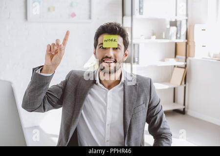 Imprenditore allegro rivolta verso l'alto e seduta con adesivo di carta sulla fronte con la parola lavoro in ufficio Foto Stock