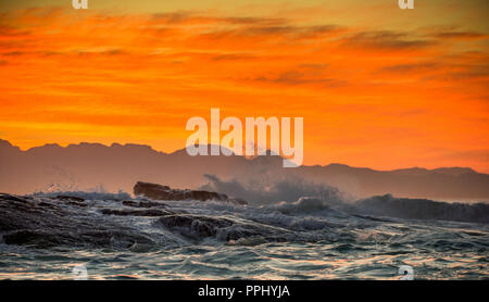 Seascape. Le nuvole in una red dawn sky, onde si infrangono con spruzzi contro sassi, sagome delle montagne all'orizzonte. False Bay. Sud Africa. Foto Stock