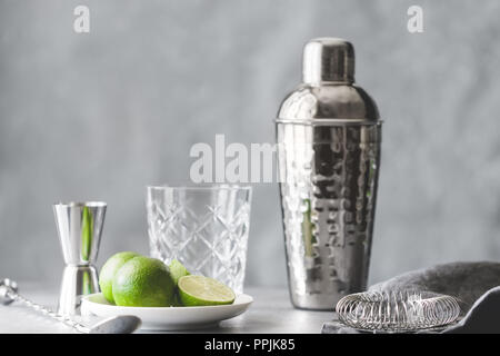 Shaker, filtro, cucchiaio da bar, vetreria, misurare e lime fresco su un tavolo. Il concetto di preparazione di cocktail e bevande alcoliche. Foto Stock