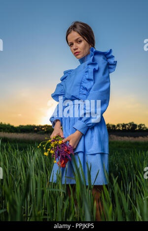 Giovane donna in blu e bianca a strisce abito è in possesso di un bouquet di fiori d'estate con due mani mentre in piedi nel campo di grano verde a spri Foto Stock