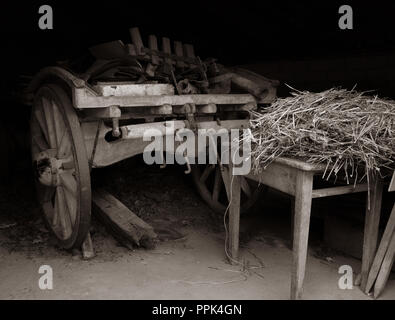 Tonalità seppia vintage a due ruote, carrello con ruote in legno, dietro un tavolo di legno e rabboccato con paglia in un vecchio fienile. Foto Stock