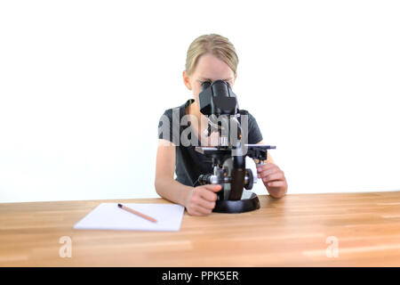 Un 9-anno-vecchia ragazza guarda in un oculare di un microscopio e rende più nitide le immagini. Lei può scrivere le sue osservazioni su una nota con una matita. Isolat Foto Stock