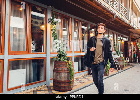 Giovane uomo con la chitarra acustica in caso a camminare su una strada di città per cafe. Hipster studente di college guy trasportare uno strumento musicale Foto Stock
