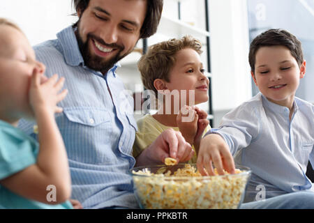 Uomo sorridente e un po' di figli mangiare popcorn durante la visione di un film insieme a casa Foto Stock