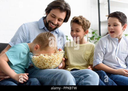 Uomo sorridente e un po' di figli mangiare popcorn durante la visione di un film insieme a casa Foto Stock