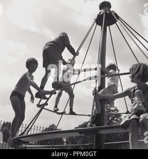 Anni sessanta, storico, giovani figli al di fuori di un campo giochi appesi su traverse in un emozionante di metallo o di oscillazione merry-go-round ride, noto come "streghe hat", Inghilterra, Regno Unito. Nel decennio più tardi, sono stati rimossi da campi da gioco a causa di preoccupazione per la salute e la sicurezza sui bambini di cadere. Foto Stock