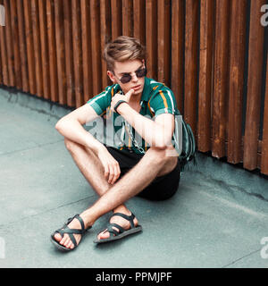 Bel uomo elegante con eleganti occhiali da sole in una camicia estiva con pantaloncini corti e un portamonete seduta vicino alla parete in legno Foto Stock