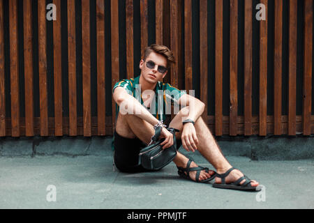 Moda giovane modello Uomo con occhiali da sole in una spiaggia shirt con pantaloncini nero seduto vicino a una parete in legno Foto Stock