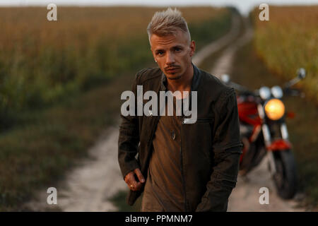 Bello ed elegante modello uomo in una camicia di cachi con una t-shirt in prossimità di un motociclo in un cornfield alla sera. Foto Stock