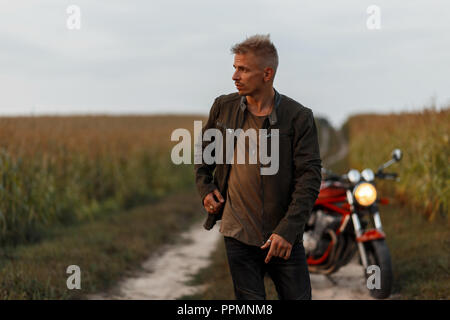 Elegante bel giovane modello uomo in una camicia cachi e t-shirt con un motociclo viaggiare sulla natura in un cornfield Foto Stock