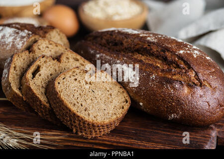 Due forme di pane di segale pane casereccio, grigio di un tovagliolo di lino, farina di grano tenero e di grano in ciotole di legno, uova e spikelets al buio su un sfondo di legno Foto Stock