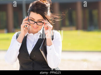 Business Woman dimensione plus si erge sullo sfondo di un edificio per uffici con una pastiglia in mano. La signora sta parlando sul suo telefono cellulare Foto Stock