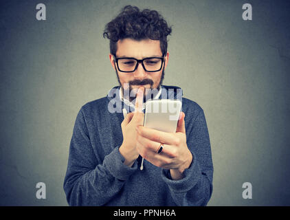 Uomo perplesso pensando cosa rispondere al messaggio di testo ricevuto sul telefono cellulare isolata sul muro grigio Sfondo. Faccia reazione di espressione del linguaggio del corpo Foto Stock