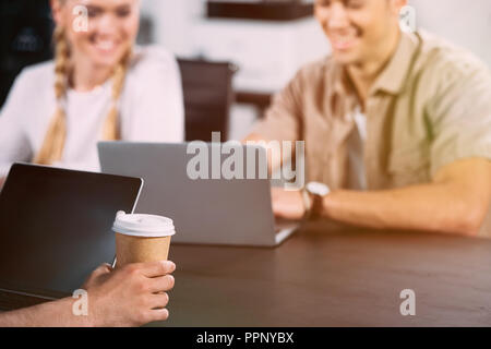 Ritagliato shot di imprenditore seduta a tavola con il computer portatile e la carta tazza di caffè mentre i suoi colleghi sorridente dietro di lavoro Foto Stock