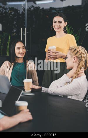 Multiculturale di business partner con bicchieri di carta di caffè avente incontro a tavola con i notebook in ufficio moderno Foto Stock