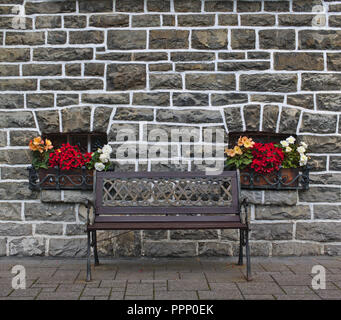 Panca in legno nella parte anteriore del grigio vecchio muro di pietra con due piccole finestre decorate con fiori colorati Foto Stock