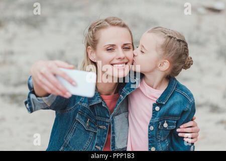 Adorabile figlia kissing madre mentre lei prendendo selfie sullo smartphone Foto Stock