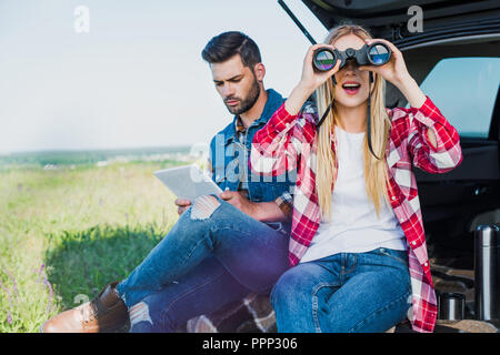 Turista femminile guardando attraverso il binocolo mentre il suo fidanzato con tavoletta digitale sul baule auto in campo rurale Foto Stock