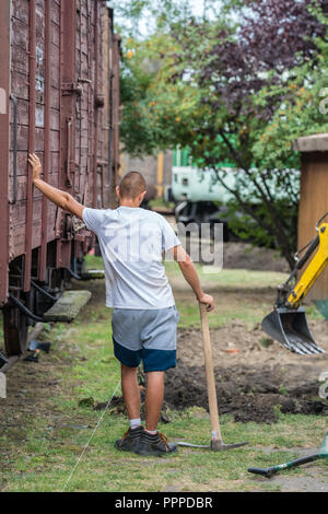 Jaworzyna slaska, Polonia - Agosto 2018 : giovane uomo in piedi da un vecchio treno in legno carrello guardando digger al lavoro Foto Stock