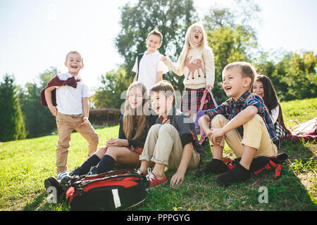 Un gruppo di sorridenti i bambini della scuola e in età prescolare sono seduti sul prato verde nel parco. L'infanzia, moda per bambini, scuola, educazione, amici, lifestyle, tempo libero, scolari concept Foto Stock