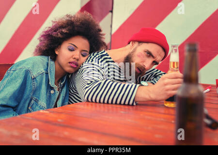 Uomo ubriaco guardando la bottiglia di birra e la sua ragazza dorme Foto Stock