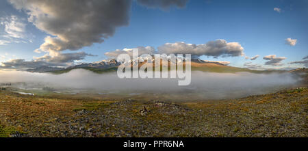 Panorama sorprendente con una verde vallata, un serpeggiante fiume, laghi e montagne coperte di neve e la foresta e la nebbia contro uno sfondo di belle nuvole Foto Stock