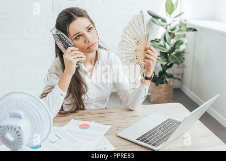 Imprenditrice stessa di raffreddamento con ventola elettrica, la ventola a mano e una bottiglia di acqua al posto di lavoro con i documenti e il computer portatile Foto Stock