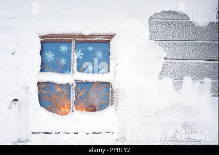 Cottage ricoperti di neve in inverno. Lume di candela incandescente attraverso frosty finestra. Foto Stock