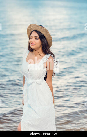 Bella ragazza felice nel cappello di paglia e abito bianco a piedi vicino al mare Foto Stock