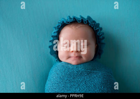 Un sorridente, tre settimane di età, i neonati bambina indossa un cofano e preinstallato in un blu turchese swaddle. Foto Stock