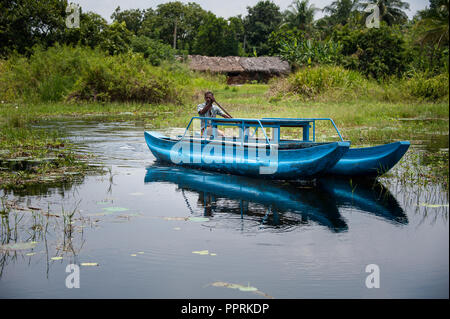 Sigiriya, Sri Lanka - Marzo 2018: un uomo locale pagaie un turchese canoe su di un lago calmo con lussureggiante giungla verde villaggio dello sfondo. Foto Stock