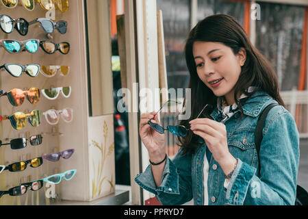 Bella asiatica è felicemente cercando occhiali da sole presso il fornitore di occhiali da sole, vuole scegliere il più adatto occhiali per lei Foto Stock