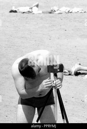 Un uomo si regola una telecamera su un treppiede mentre al beach, ca. 1955. Foto Stock