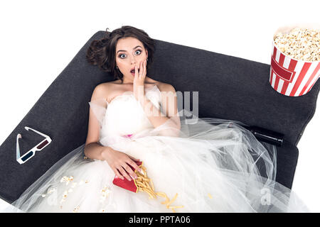 Angolo di alta vista scioccato giovane sposa in abito da sposa seduto sul divano con il cibo spazzatura isolato su bianco Foto Stock