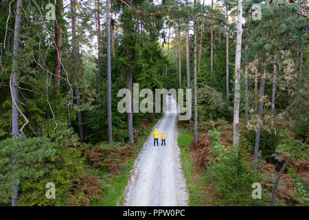 La madre e il figlio che indossa giacche gialle in piedi nel centro della strada di ghiaia in una foresta da un drone su un pomeriggio autunnale Foto Stock