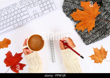 Caffè con marshmallow in mani su un desktop bianco accanto a un blocco vuoto e tastiera, foglie di autunno. Caduta dell'umore. Vista superiore, disposizione piatta. Copiare la spa Foto Stock