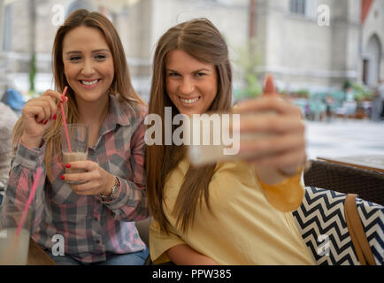 Due allegri allegro ragazze prendendo un selfie mentre è seduto presso il cafe. Felice giovani donne divertimento in coffee shop. Migliori amici prendendo un selfie con smart Foto Stock