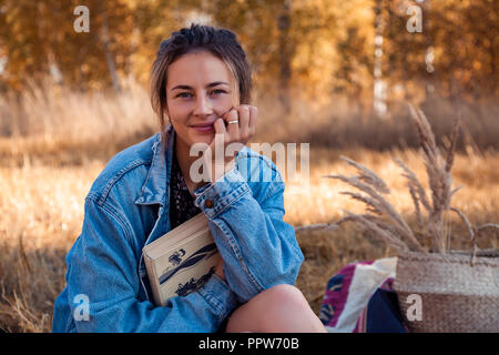 Pic-nic all'aria fresca: una giovane donna in una camicia di jeans e abiti gode di natura, pone e mantiene un libro sul retro di uno sfondo del campo di autunno l Foto Stock