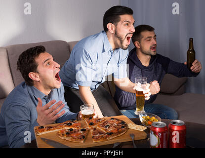 Esaltato amici maschi guardando la tv insieme a casa, godendo di birra e pizza Foto Stock