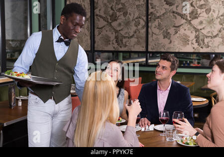 Sorridente African American cameriere che serve pasti al cordiale compagnia nel ristorante accogliente Foto Stock