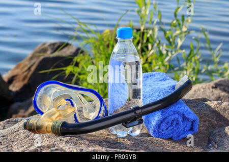 Bottiglia con acqua, asciugamano, maschera per immersioni subacquee e snorkeling tubo su roccia con la riva del mare sullo sfondo. Estate, uno stile di vita attivo Foto Stock