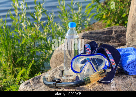 Bottiglia con acqua, asciugamano, flip-flop, maschera per immersioni subacquee e snorkeling tubo giacciono su pietre con riva del mare sullo sfondo. Estate, uno stile di vita attivo Foto Stock