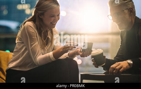 Donna sorridente con matura collega seduto nella lobby di Office utilizzando il telefono cellulare durante la pausa caffè. Due persone sedute in ufficio looki lounge Foto Stock